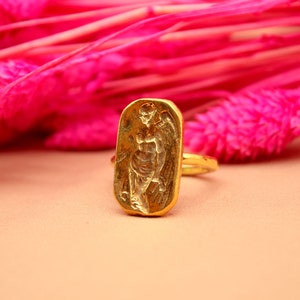 May include: A gold ring with a rectangular design featuring a raised image of a winged figure. The ring is set against a pink background.