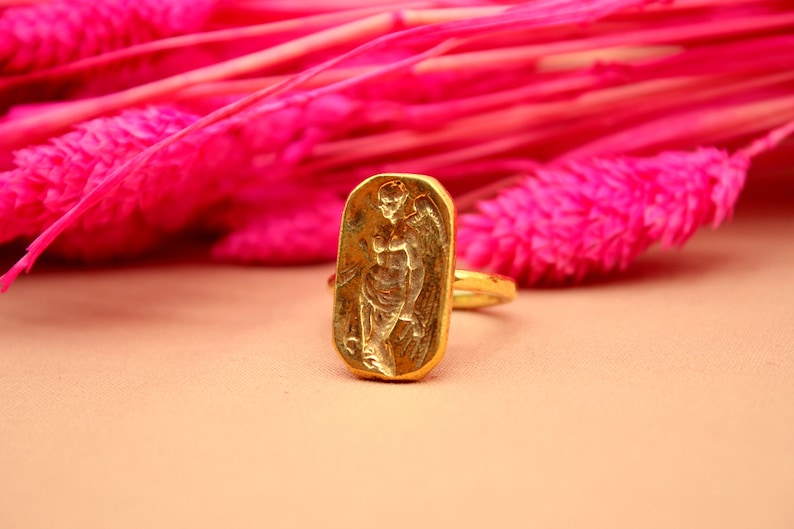 May include: A gold ring with a rectangular design featuring a raised image of a winged figure. The ring is set against a pink background.