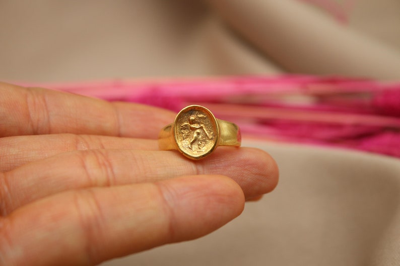 May include: A gold ring with a raised oval design featuring a detailed figure. The ring is worn on a finger.