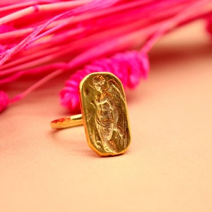 May include: A gold ring with a rectangular design featuring an embossed image of an angel with wings.