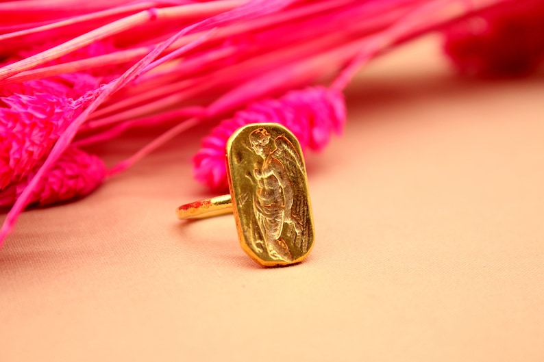 May include: A gold ring with a rectangular design featuring an embossed image of an angel with wings.