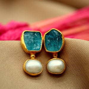 May include: A pair of gold earrings with blue gemstone and pearl accents. The earrings feature a unique, irregular shaped blue gemstone and a round pearl.
