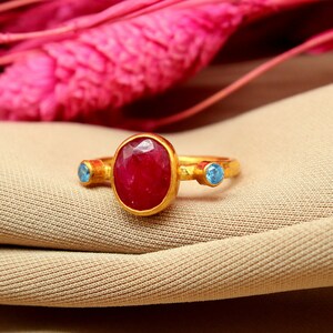 K&ouml;nnte beinhalten: Ein goldener Ring mit einem gro&szlig;en ovalen Rubin-Edelstein und zwei kleineren blauen Edelsteinen auf jeder Seite.