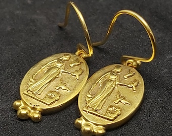 May include: Gold tone earrings with a detailed oval pendant featuring a woman holding a bird. The pendant is attached to a gold tone hook.