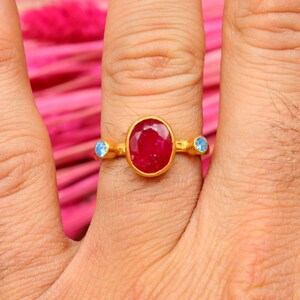 K&ouml;nnte beinhalten: Ein goldener Ring mit einem gro&szlig;en ovalen roten Edelstein und zwei kleineren blauen Edelsteinen an beiden Seiten.
