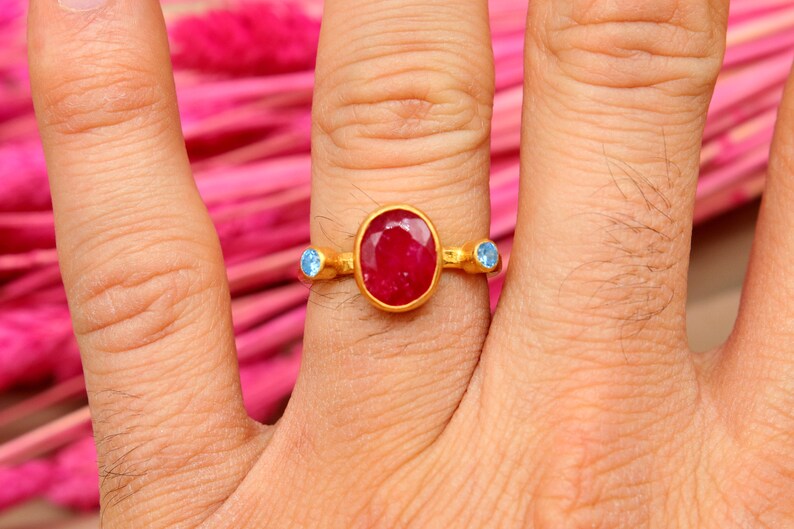 K&ouml;nnte beinhalten: Ein goldener Ring mit einem gro&szlig;en ovalen roten Edelstein und zwei kleineren blauen Edelsteinen an beiden Seiten.