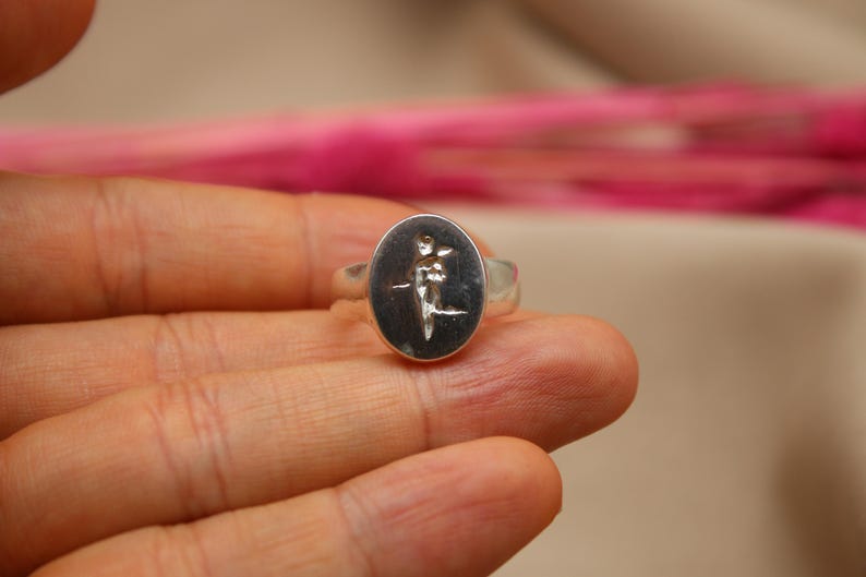 以下が含まれることがあります： 花を持っている人の詳細なデザインが施された、楕円形のセッティングのシルバーリングです。