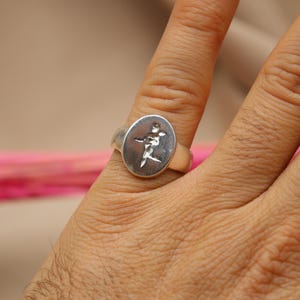 以下が含まれることがあります： スタイリッシュな人物が描かれた楕円形のデザインのシルバーリング。指に着用されています。