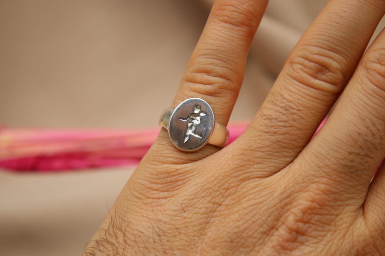 以下が含まれることがあります： スタイリッシュな人物が描かれた楕円形のデザインのシルバーリング。指に着用されています。