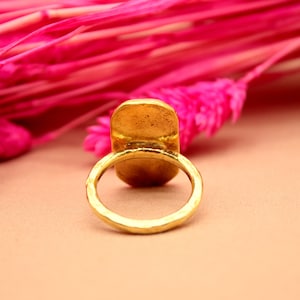 May include: A gold-toned ring with a large oval-shaped setting. The ring is sitting on a light brown surface.