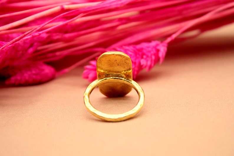 May include: A gold-toned ring with a large oval-shaped setting. The ring is sitting on a light brown surface.