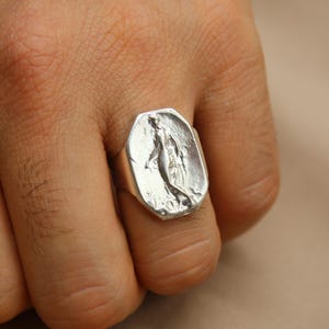 May include: A silver ring with a rectangular face featuring a sculpted figure. The ring is worn on a finger, showcasing the detailed design and metallic sheen. The ring's design is a unique piece of jewelry.