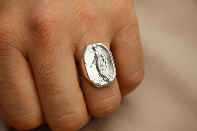 May include: A silver ring with a rectangular face featuring a sculpted figure. The ring is worn on a finger, showcasing the detailed design and metallic sheen. The ring's design is a unique piece of jewelry.