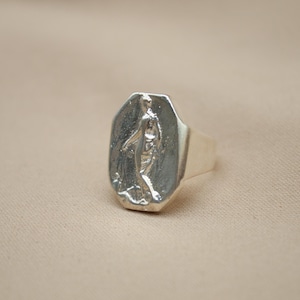 May include: A silver ring with a rectangular face featuring a detailed relief of a figure. The ring has a polished finish and a thick band. The design is set against a neutral background.