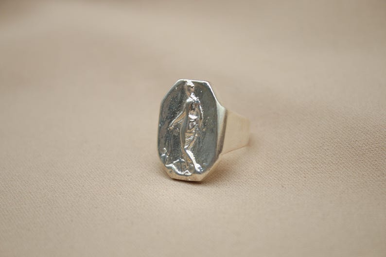 May include: A silver ring with a rectangular face featuring a detailed relief of a figure. The ring has a polished finish and a thick band. The design is set against a neutral background.