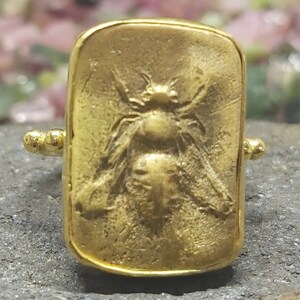 May include: A gold-toned ring with a rectangular design featuring a detailed embossed image of a bee.