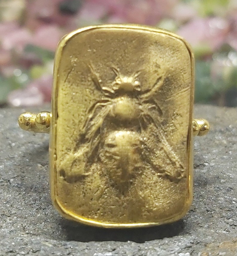 May include: A gold-toned ring with a rectangular design featuring a detailed embossed image of a bee.