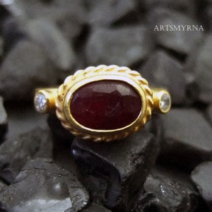 K&ouml;nnte beinhalten: Ein goldener Ring mit einem gro&szlig;en ovalen Rubin-Edelstein und zwei kleineren Diamanten. Der Ring ist in einem seilartigen Design gefasst.