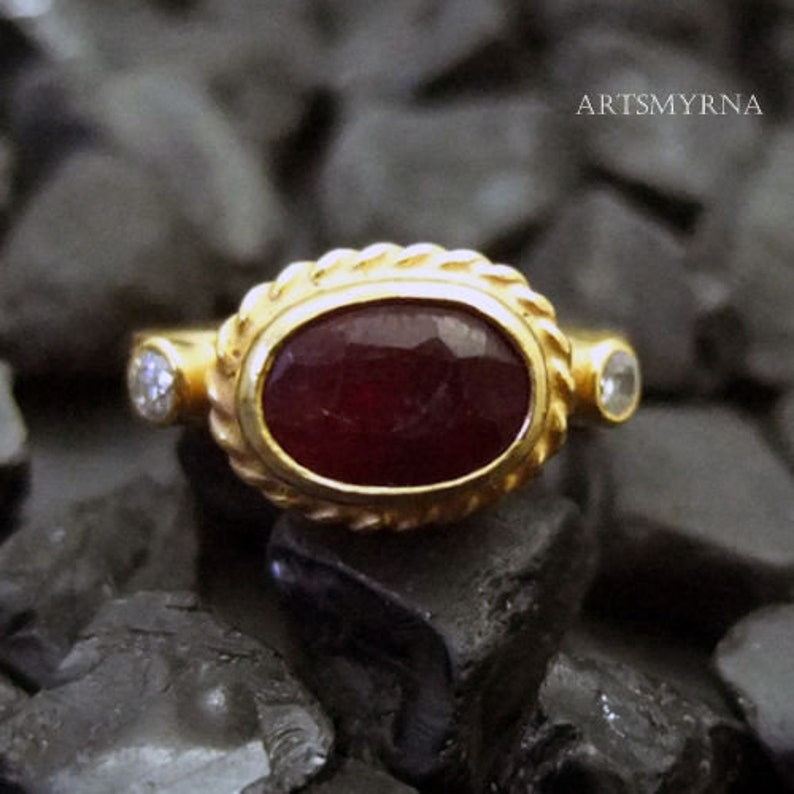 K&ouml;nnte beinhalten: Ein goldener Ring mit einem gro&szlig;en ovalen Rubin-Edelstein und zwei kleineren Diamanten. Der Ring ist in einem seilartigen Design gefasst.