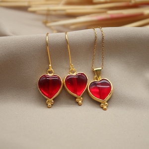 May include: A set of gold heart-shaped earrings and a necklace with red glass hearts. The jewellery is on a beige fabric background.