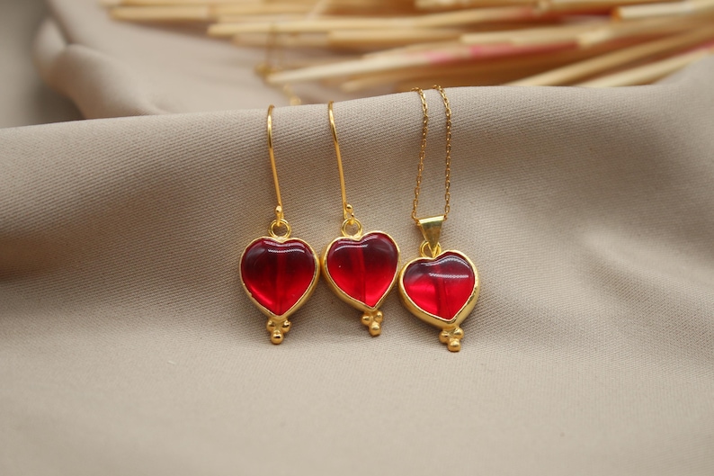 May include: A set of gold heart-shaped earrings and a necklace with red glass hearts. The jewellery is on a beige fabric background.