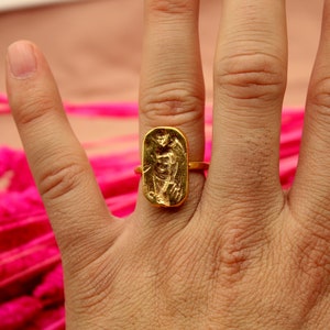 May include: A gold ring with a rectangular design featuring a raised image of a person. The ring is worn on a person's finger.