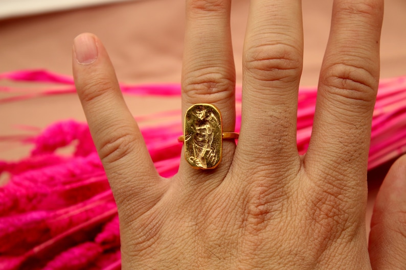May include: A gold ring with a rectangular design featuring a raised image of a person. The ring is worn on a person's finger.