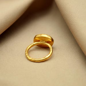 May include: A gold ring with a large oval-shaped gemstone. The ring is set on a beige fabric background.