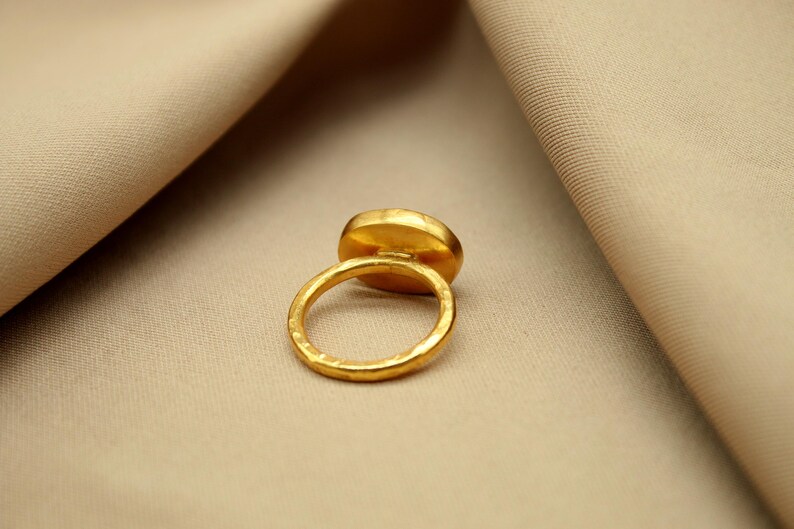 May include: A gold ring with a large oval-shaped gemstone. The ring is set on a beige fabric background.