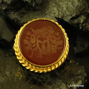 May include: A round, antique-style brooch with a gold-colored, twisted rope border. The center features a reddish-brown stone with a detailed, engraved design. The brooch is set against a dark, textured background.