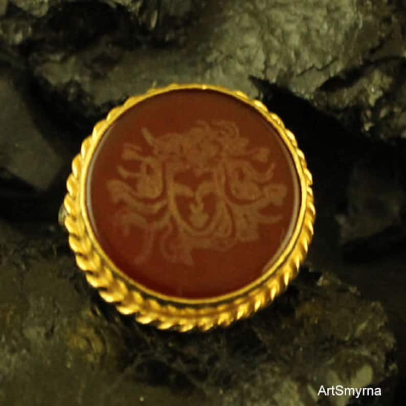May include: A round, antique-style brooch with a gold-colored, twisted rope border. The center features a reddish-brown stone with a detailed, engraved design. The brooch is set against a dark, textured background.
