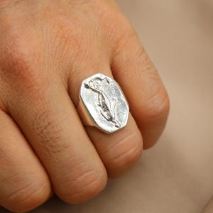 May include: A silver ring with a rectangular face featuring a floral design. The ring is worn on a finger, showcasing its polished metal and detailed engraving. The ring's design is a stylized flower.