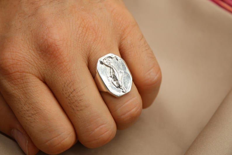 May include: A silver ring with a rectangular face featuring a floral design. The ring is worn on a finger, showcasing its polished metal and detailed engraving. The ring's design is a stylized flower.