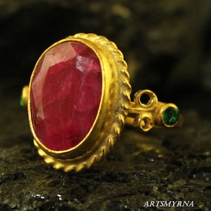 May include: A gold ring featuring a large, oval ruby gemstone set in a twisted rope design. The ring has two small green gemstones on the band.