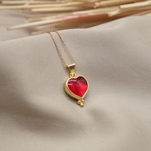 May include: A gold chain necklace with a red heart pendant. The heart is detailed with gold accents and has a slightly textured surface.