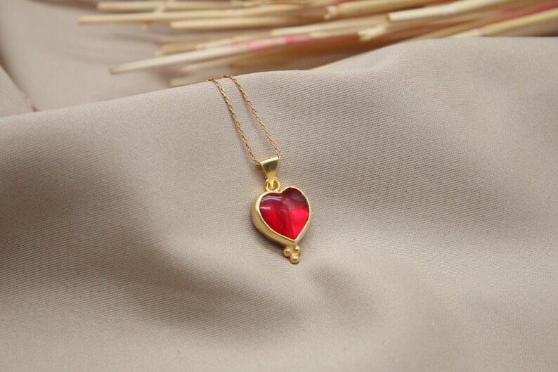 May include: A gold chain necklace with a red heart pendant. The heart is detailed with gold accents and has a slightly textured surface.