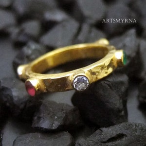 Op de afbeelding: Gouden ring met een gehammerde textuur en vijf edelstenen. De edelstenen zijn een heldere diamant, een rode robijn en drie groene smaragden.