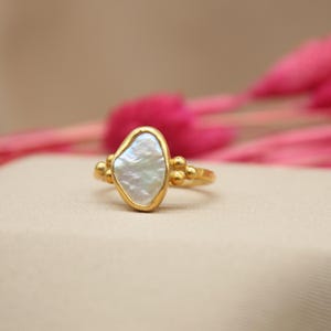 May include: A gold ring with a white, irregular shaped pearl-like stone set in the center. The ring has small gold beads on either side of the stone.