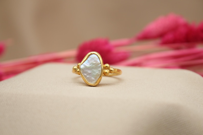 May include: A gold ring with a white, irregular shaped pearl-like stone set in the center. The ring has small gold beads on either side of the stone.