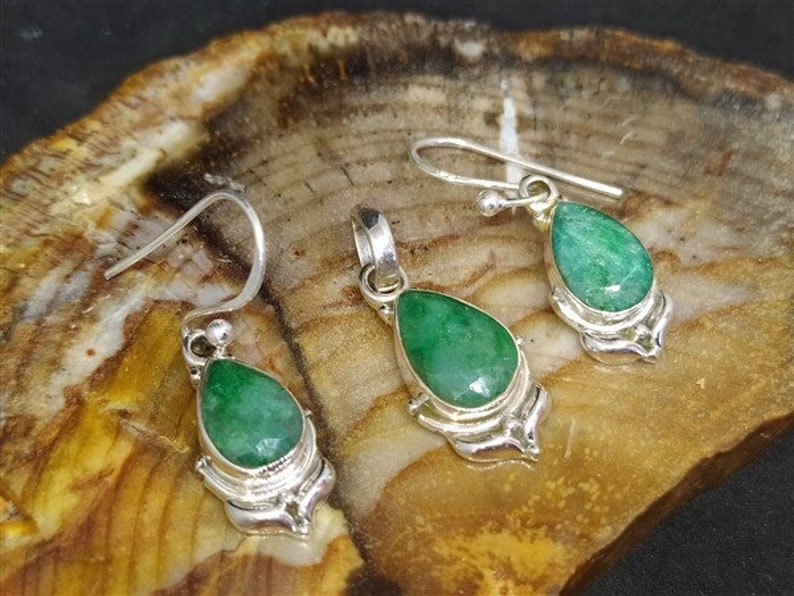 Pu&ograve; includere: Un set di gioielli con un ciondolo e orecchini abbinati. Ogni pezzo ha una pietra preziosa verde sfaccettata a forma di goccia incastonata in metallo color argento. Gli orecchini hanno un design a gancio e il ciondolo ha un anello per una catena.