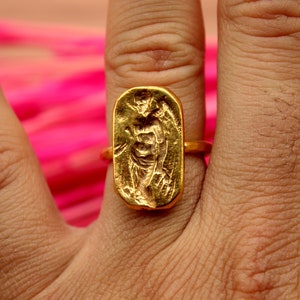 May include: A gold ring with a rectangular design featuring a raised image of a person. The ring is worn on a person's finger.