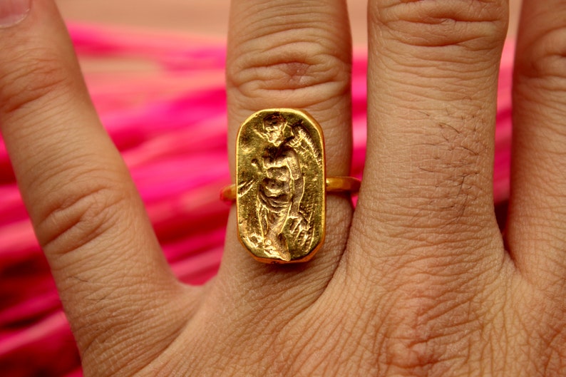 May include: A gold ring with a rectangular design featuring a raised image of a person. The ring is worn on a person's finger.