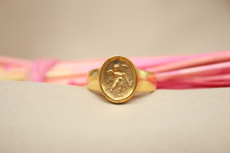 May include: A gold ring with an oval-shaped design featuring a raised image of a person in a running pose.