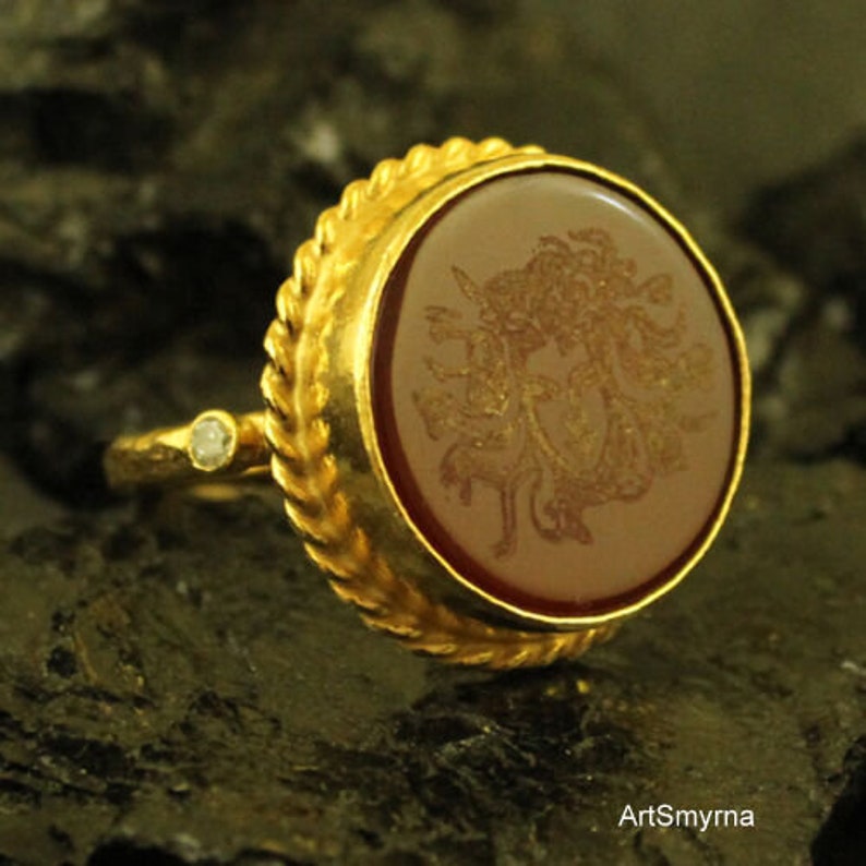 May include: A gold ring with a large, oval, reddish-brown stone featuring a detailed engraved design. The ring has a textured, braided band and a small, clear gemstone on the side. The ring is set against a dark, textured background.