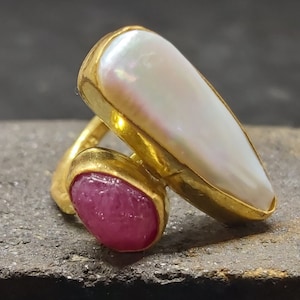 May include: A gold ring with a large white pearl and a pink gemstone. The ring is set on a rough, gray surface.