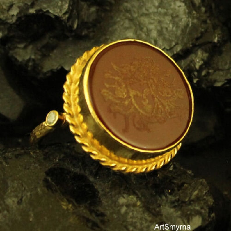 May include: A gold ring with a large, round, reddish-brown stone. The stone has a detailed, golden design. The ring has a decorative, gold leaf-like border around the stone and a small, clear stone on the band.