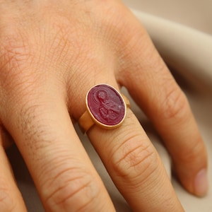 May include: A gold ring with a red oval gemstone featuring a carved design. The ring is worn on a finger.
