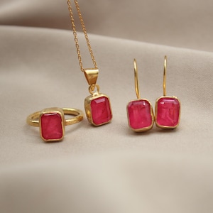 May include: A gold jewelry set with a pendant, earrings, and a ring. Each piece features a square-shaped pink gemstone.