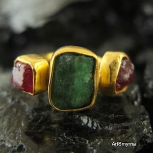 Op de afbeelding: Een gouden ring met drie edelstenen: een grote groene smaragd in het midden en twee kleinere rode robijnen aan weerszijden.