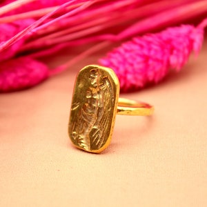 May include: A gold ring with a rectangular design featuring an embossed image of a winged figure. The ring is set against a pink background.
